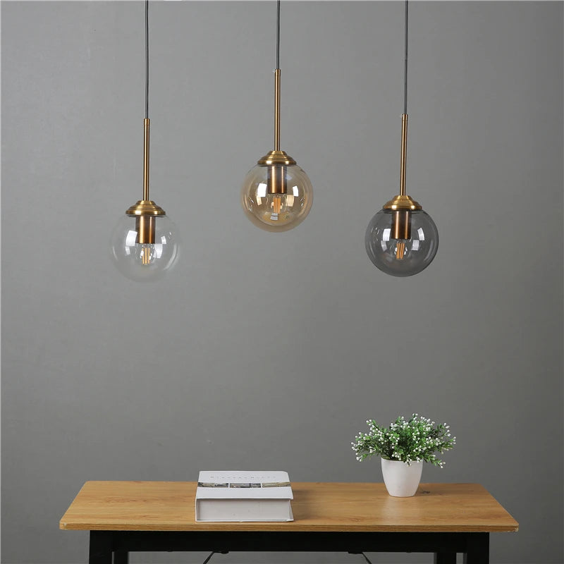 Three pendant lights with glass shades hanging above a wooden table with books and a plant.