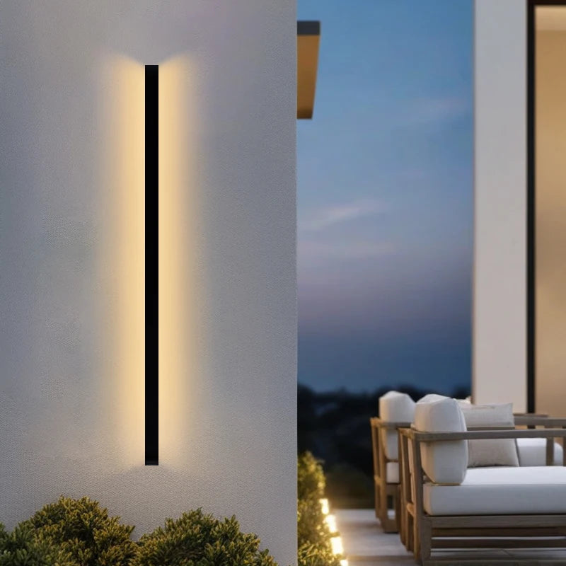 Modern outdoor wall light fixture on a building exterior with a patio and seating area in the background.