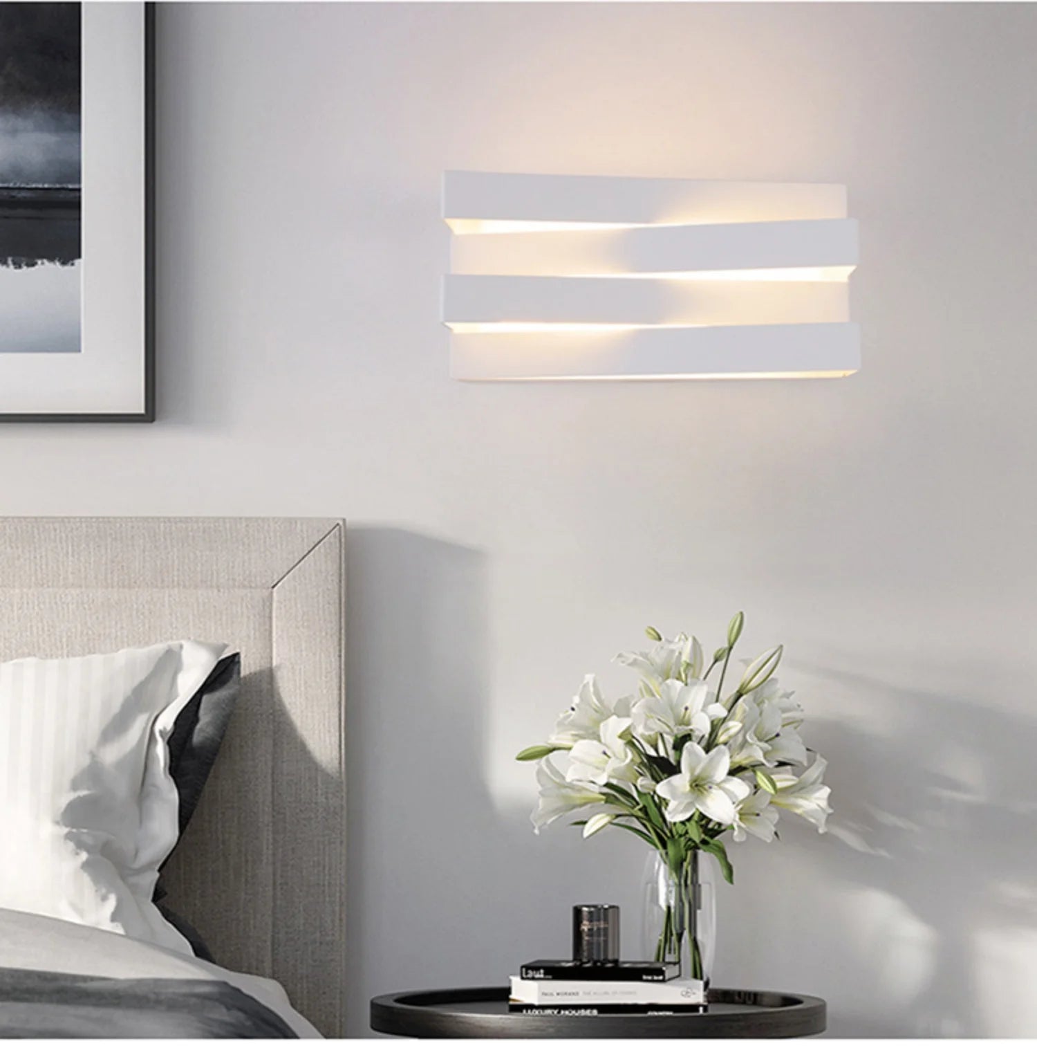 Modern wall sconce above a bed with a vase of white flowers on a nightstand.