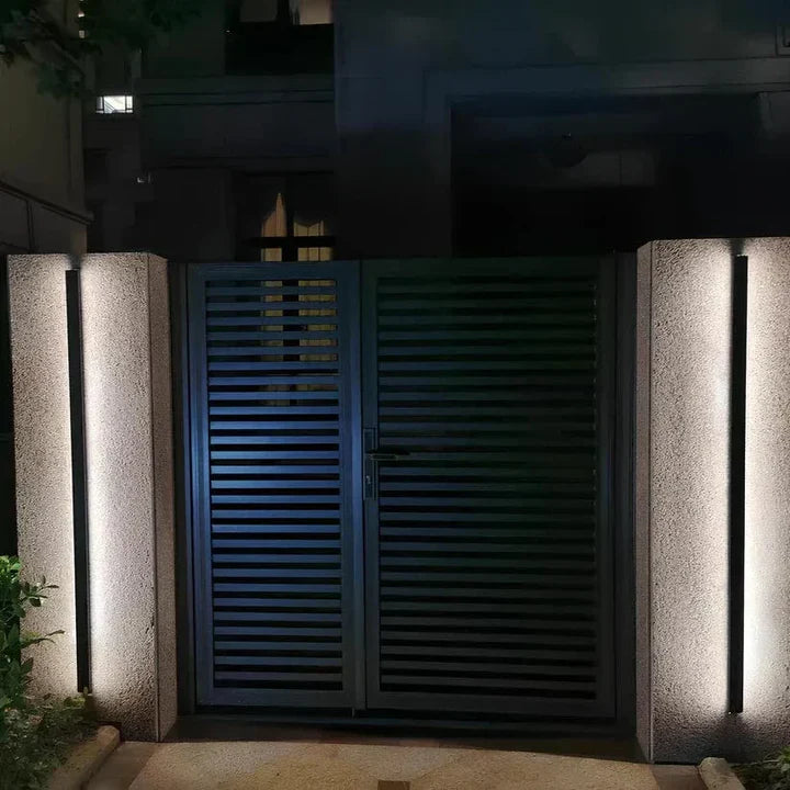 Modern gate with stone pillars illuminated by outdoor wall lights at night.